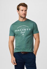 Homme portant un T-shirt vert « Hackett Sport » et un pantalon bleu marine, debout avec une main en poche devant un fond clair uni.
