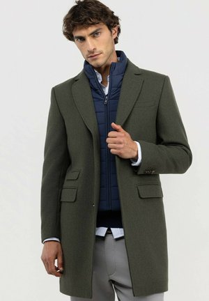 Hombre con un abrigo largo verde oscuro sobre una chaqueta azul acolchada y pantalones gris claro, ajustándose el abrigo con una mano.