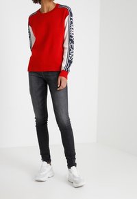 Czerwony sweter z białymi i granatowymi akcentami oraz napisem "TOMMY JEANS" na rękawach, w połączeniu z ciemnymi dżinsami i białymi trampkami.