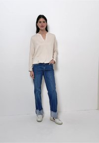 Vrouw staande tegen een witte muur, gekleed in een beige blouse met lange mouwen, blauwe omgeslagen spijkerbroek en witte sneakers, met ontspannen handen in de zak.