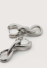 Λουστραρισμένα σκουλαρίκια σε ασημί τόνο με στριφογυριστό σχέδιο άπειρου και κούμπωμα με καρφί, σε απλό λευκό φόντο.