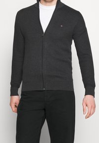 Cardigan grigio con zip realizzato in tessuto a maglia testurizzato, dotato di colletto alto e polsini a coste, con un piccolo dettaglio del logo sul petto.