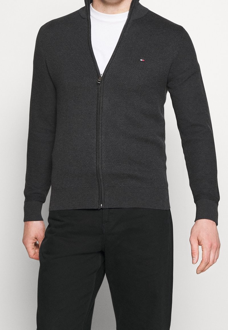 Cardigan grigio con zip realizzato in tessuto a maglia testurizzato, dotato di colletto alto e polsini a coste, con un piccolo dettaglio del logo sul petto.