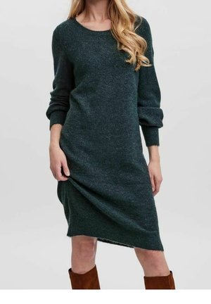 Nainen, joka pukeutunut tummanvihreään pitkähihaiseen neulemekkoon, jossa on resorit hihansuissa ja helmassa, yhdistettynä ruskeisiin polvenkorkuisiin saappaisiin.