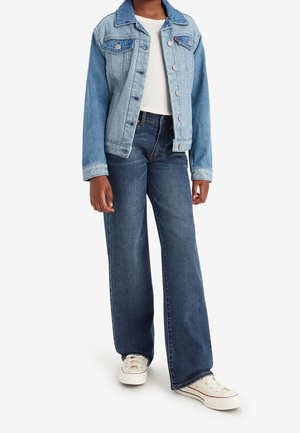 Jasnoniebieska dżinsowa kurtka zapinana na guziki, z dwoma kieszeniami na piersi, oraz szerokie ciemnoniebieskie dżinsy, zestawione z białymi sneakersami z płótna.