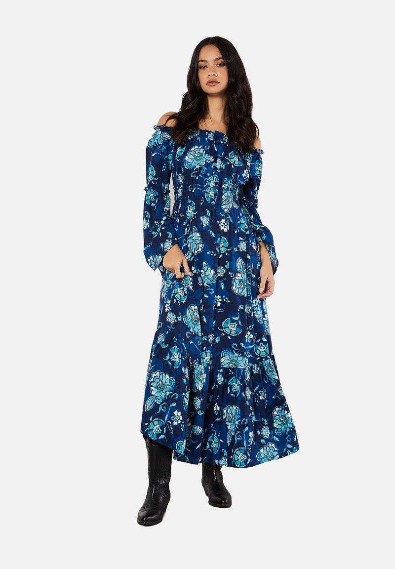 Blommig off-shoulder maxiklänning i marinblått med blå och turkosa mönster. Har lång ärm, elastisk midja och volangkant. Matchas med svarta stövlar.