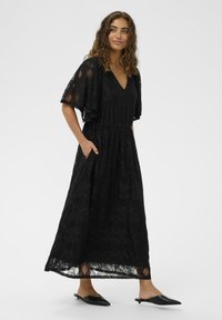 Fekete csipke maxi ruha V-nyakkal, fodros ujjakkal és szűkített derékkal. Texturált mintázattal és oldalsó zsebekkel rendelkezik.