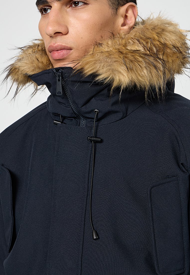 Veste navy avec une capuche en fourrure, dotée d'un col haut, de cordons de serrage réglables et de deux poches avant. Texture lisse, design utilitaire.