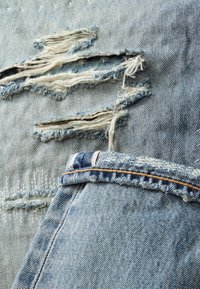 Tissu en denim bleu avec de multiples déchirures horizontales et des fils blancs effilochés, à côté d'un denim plié avec une couture visible et des surpiqûres orange.