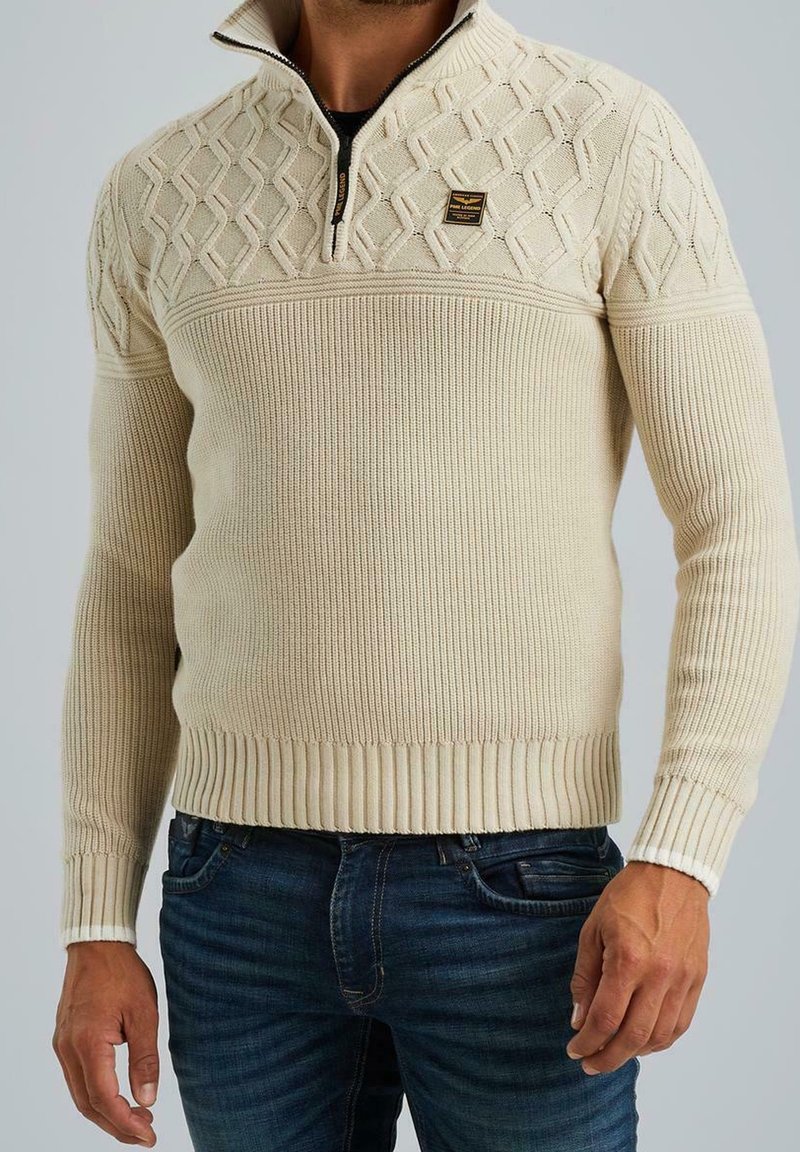Homme portant un pull beige en maille texturée avec col demi-fermeture éclair et un jean bleu foncé, debout devant un fond uni.