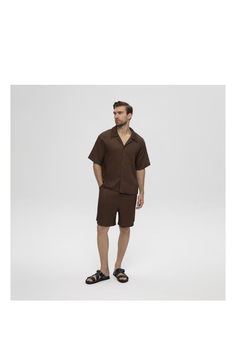 Man staat met handen in de zakken, draagt een bruin shirt met korte mouwen, bijpassende korte broek en zwarte sandalen tegen een effen achtergrond.