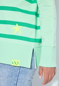 Camisa de manga larga verde claro con rayas verde oscuro, estrella amarilla y símbolos de infinito, usada con jeans azul claro, mano relajada al costado.