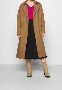 Kobieta w brązowym płaszczu typu trench, jasnoróżowej bluzce z dekoltem w serek, czarnej plisowanej spódnicy i beżowych botkach do połowy łydki na jednolitym tle.