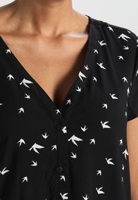 Blusa nera con scollatura a V, maniche corte e stampa di uccelli bianchi. Presenta bottoni frontali e un tessuto leggero e strutturato.