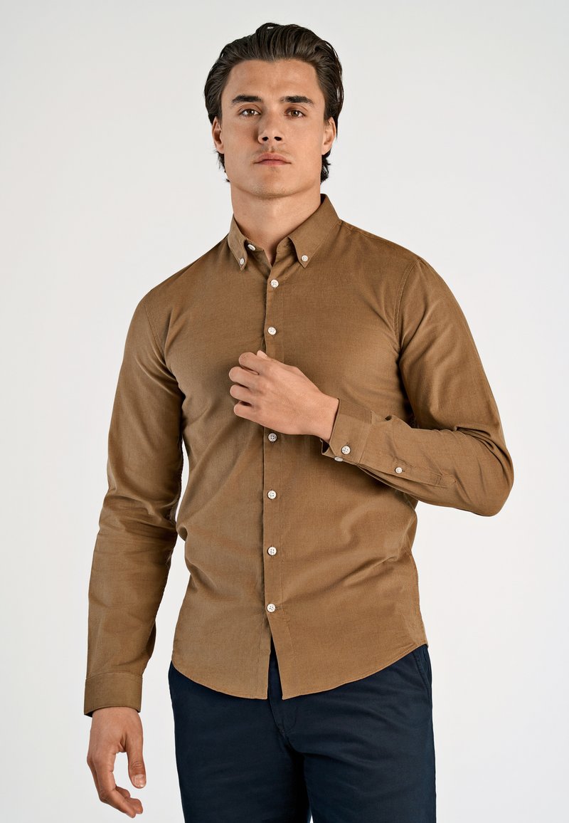 Camicia marrone con bottoni, a maniche lunghe, realizzata in tessuto testurizzato. Presenta bottoni bianchi e una vestibilità comoda. Indossata con pantaloni scuri.