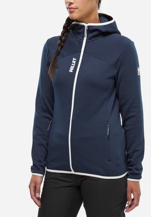 Woman wearing a navy blue zip-up hoodie with white trim, white zipper, and Millet logo on chest and sleeve, standing with her hands relaxed.