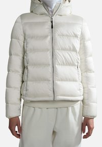 Biała kurtka puchowa z kapturem, krótka, o pikowanej fakturze, z zamkiem błyskawicznym z przodu i elastycznymi mankietami. Noszona na dopasowanych beżowych spodniach.