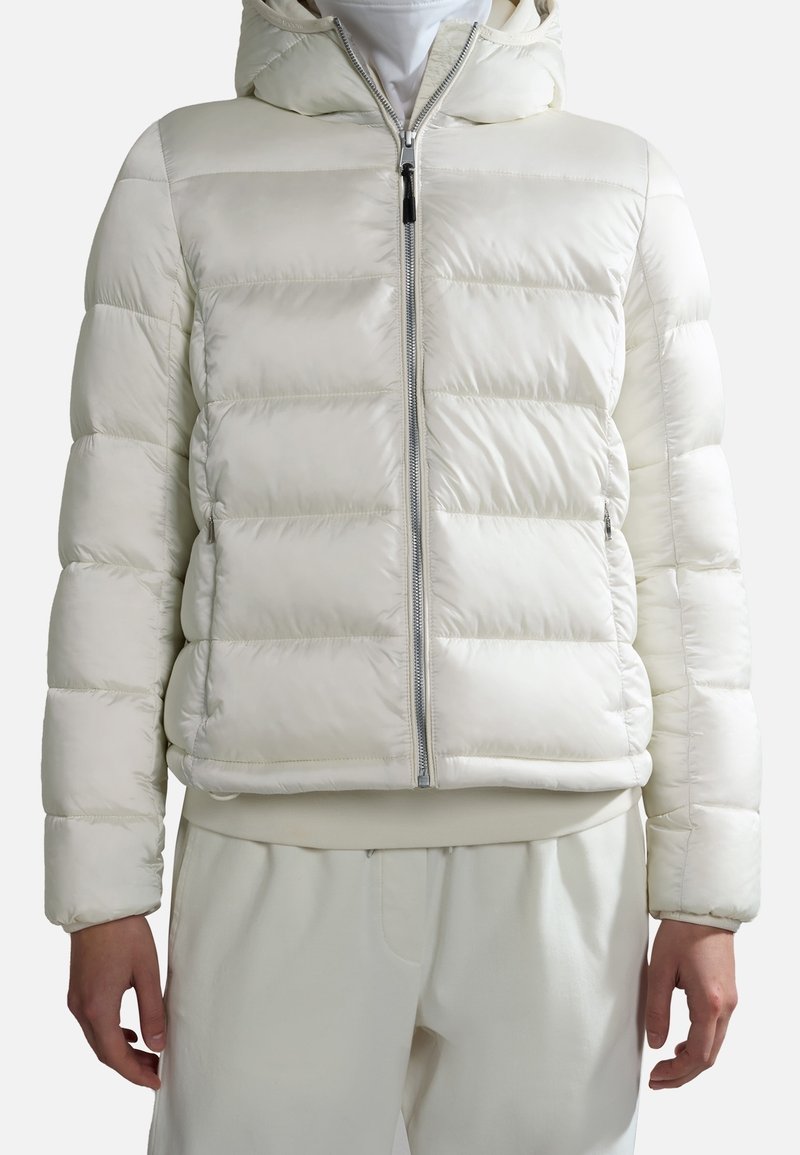 Biała kurtka puchowa z kapturem, krótka, o pikowanej fakturze, z zamkiem błyskawicznym z przodu i elastycznymi mankietami. Noszona na dopasowanych beżowych spodniach.