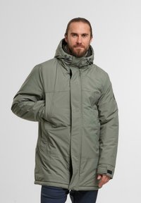 Parka imbottita in verde oliva, con colletto alto, cappuccio regolabile e tasche frontali. Tessuto liscio con cerniera nascosta.