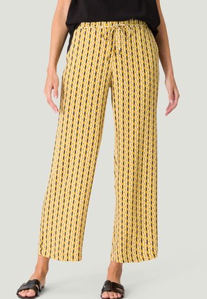 Femme portant un pantalon large jaune à motifs, un haut noir et des sandales noires, debout devant un fond clair uni.