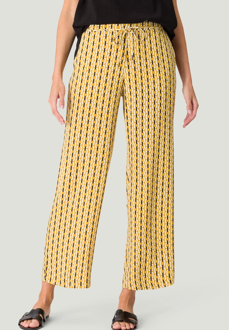 Femme portant un pantalon large jaune à motifs, un haut noir et des sandales noires, debout devant un fond clair uni.