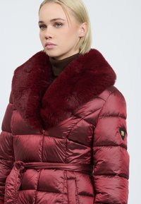 Blonde vrouw in een glanzende bordeauxgekleurde gewatteerde jas met een grote bontkraag en riem, gecombineerd met een bruine coltrui.