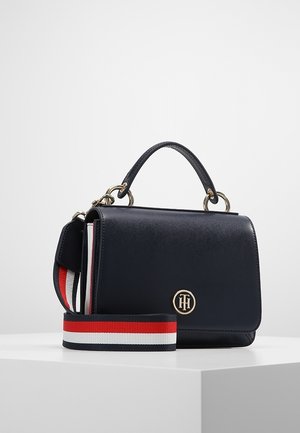 Bolso de cuero negro con asa superior, emblema de logo dorado y una correa desmontable en rayas rojas, blancas y azul marino, colocado sobre una superficie blanca.