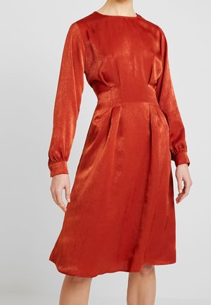 Kvinde iført en langærmet, knælængde, brændt orange satin kjole med tætsiddende talje og rund halsudskæring mod en ensfarvet baggrund.