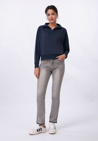 Mørkeblå pullover med kvart glidelås og ribbestrikket kant, parret med lysegrå jeans og hvite sneakers med svarte striper.