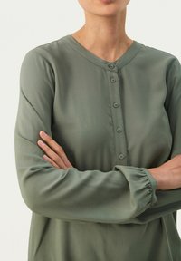 Olivengrøn, langærmet bluse i glat stof. Rund halsudskæring med fem matchende knapper og samlede mansjetter på ærmerne.