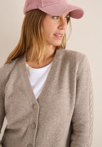Beige stickad kofta med V-ringning, som har små knappar och flätstickade detaljer på ärmarna. Matchas med en vit T-shirt och rosa keps.