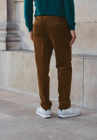 Pantaloni din corduroy maron cu croială dreaptă și două buzunare la spate, purtați cu adidași gri deschis cu accente verzi, pe un fundal neutru.