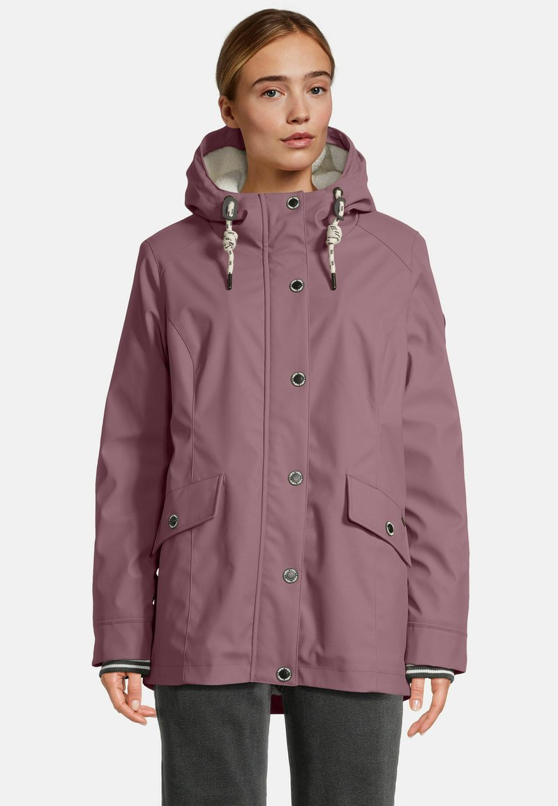 Frau in einer staubigen rosafarbenen Jacke mit Kapuze, Frontknöpfen, Kordelzugkapuze, Klappentaschen und grauen Hosen, die vor einem schlichten Hintergrund steht.