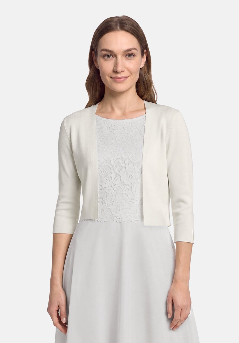 Vrouw met lang bruin haar draagt een witte open cardigan met driekwart mouwen over een witte kanten jurk, staand tegen een lichtgrijze achtergrond.