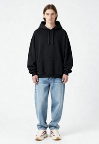 Felpa nera oversize con cappuccio e lacci, tasca frontale, polsini a costine, abbinata a jeans azzurri chiari e sneakers multicolori.