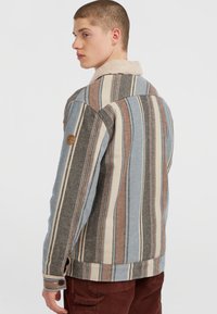Jeune homme portant une veste rayée avec des rayures verticales bleues, brunes et beiges, et un col en sherpa beige, de profil contre un fond blanc.