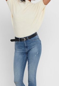 Jasno-beżowy prążkowany top zestawiony z niebieskimi skinny jeansami, z czarnym paskiem. Jeansy mają niewielkie przetarcia w okolicy kolan.