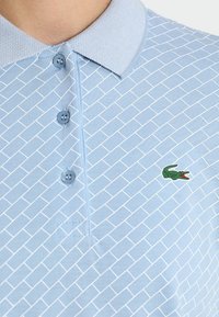 Ljusblå pikétröja med ett strukturerat geometriskt mönster, tre blå knappar och en grön krokodil-logotyp på vänster sida.