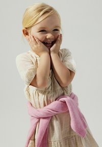 Robe beige clair avec manches bouffantes et taille froncée, accompagnée d'un pull rose pâle noué autour de la taille. Fond neutre.