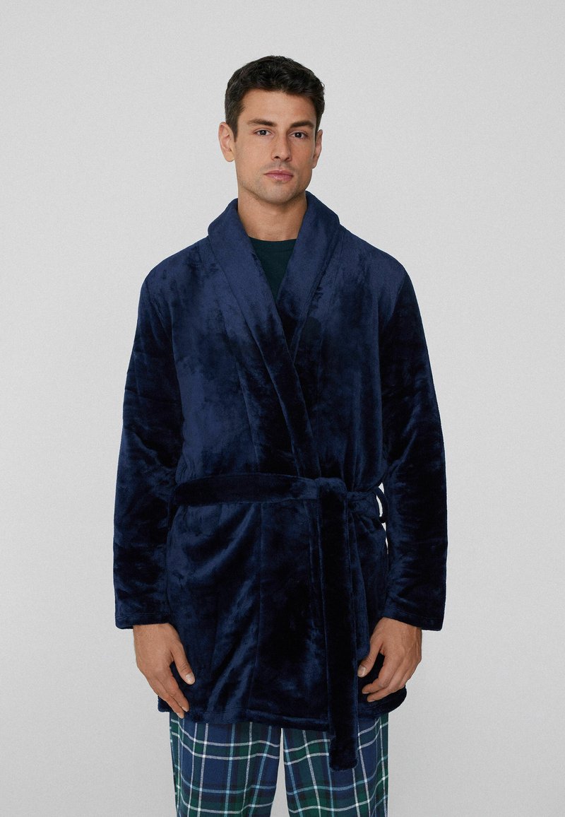 Tmavě modrý plyšový župan s šálovým límcem, páskem na zavazování v pase a dlouhými rukávy, nošený přes kárované pyžamové kalhoty v zelené a modré barvě.