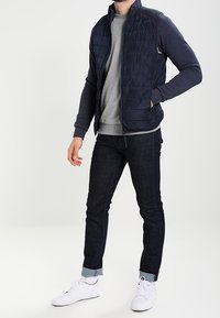 Marinblå quiltad väst med hög krage, tillsammans med en grå sweatshirt och mörka jeans. Outfitten fulländas med vita sneakers.