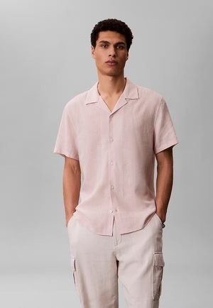 Uomo che indossa una camicia rosa chiaro a maniche corte con bottoni e pantaloni cargo di colore chiaro, in piedi davanti a uno sfondo grigio uniforme.
