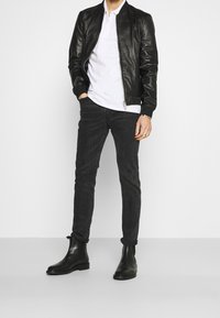 Man som bär en svart läderjacka, vit zip-up tröja, svarta skinny jeans och svarta läder ankelstövlar, står mot en enkel bakgrund.