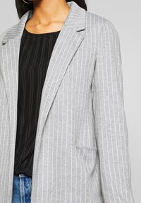 Mujer que lleva un blazer gris de rayas sobre una blusa negra con pliegues y vaqueros azules, mostrada desde el cuello hasta las caderas.