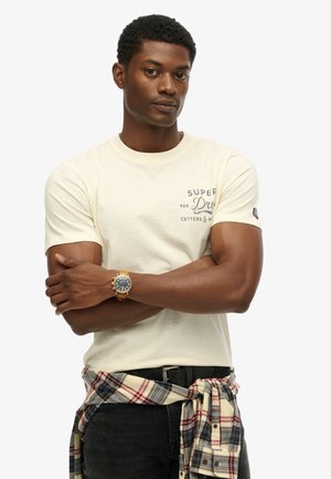 Cream short-sleeve t-shirt with logo print, paired with a plaid shirt tied at the waist. Black pants and a gold watch accentuate the look.