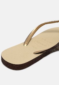 Sandală flip-flop din cauciuc aurie, cu un călcâi texturat de culoare bej. Talpa maronie cu sclipici accentuează designul. Curelele subțiri și curbe au mici embellismente.
