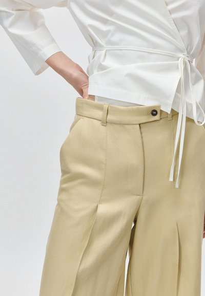 Μπεζ πλισέ παντελόνι ψηλόμεσο με κουμπί και θηλιές για ζώνη, σε συνδυασμό με ένα λευκό πουκάμισο τύπου wrap δεμένο στη μέση.