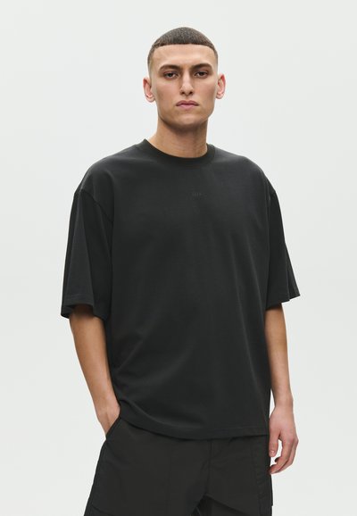Jeune homme aux cheveux courts portant un T-shirt noir uni oversize et un pantalon noir, debout avec une main dans la poche devant un fond uni.