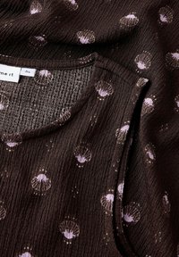 Haut en tissu texturé marron avec un col rond, comportant des motifs de coquillages violets clairs disséminés et une surface douce et froissée.