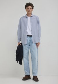 Camicia azzurra a maniche lunghe con bottoni sopra una t-shirt bianca, abbinata a jeans azzurri e scarpe in suede marroni. Maglione scuro appoggiato su un braccio.